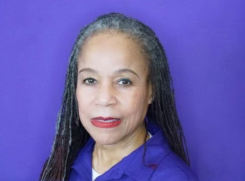A woman with gray hair is wearing a purple shirt and red lipstick.