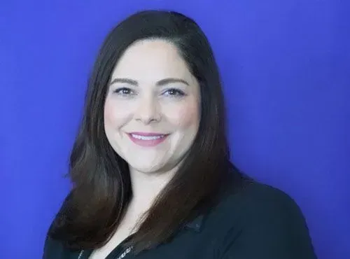 A woman in a black jacket is smiling in front of a blue background.