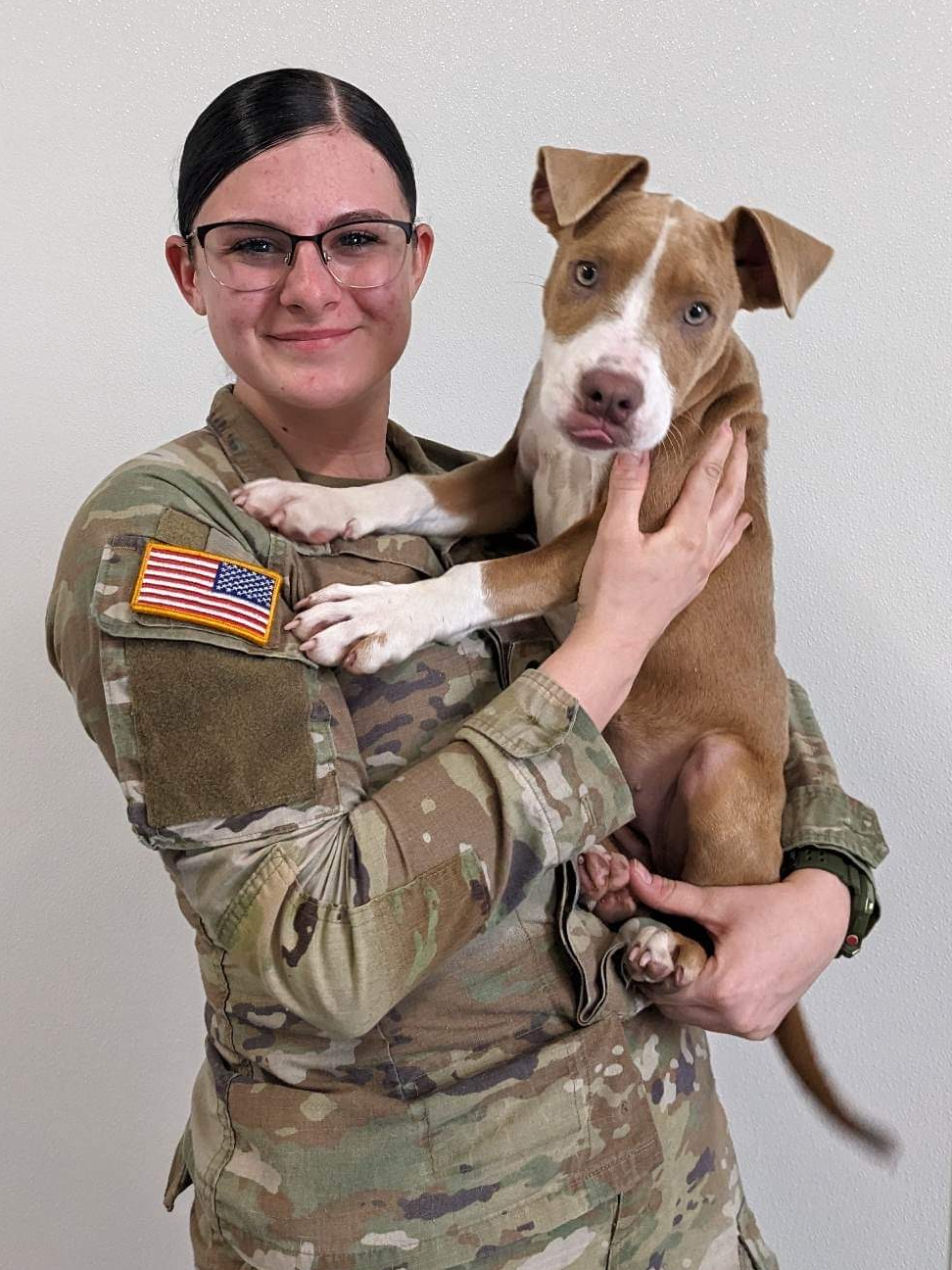 Woman in military uniform holding a brown and white puppy, both smiling. They are indoors, with a plain white background.