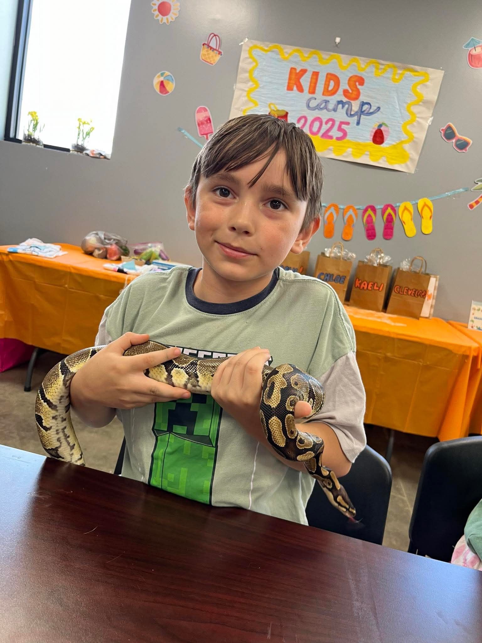 A young boy smiles, holding a patterned snake. He sits at a table in a room decorated for 