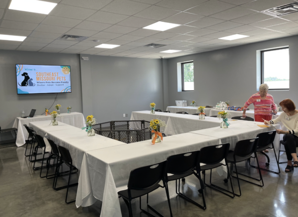 Conference room set up with a U-shaped table, featuring decorations and a screen displaying a presentation. Two women are preparing the room.