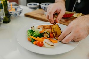 Hands plating roasted vegetables and chicken on a white dinner plate in a bright kitchen