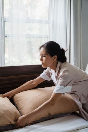 Person making a bed by a bright window, smoothing a beige blanket on a white mattress.