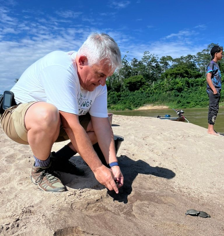Man crouching on a sandy hill, examining the ground beside a standing person under a blue sky.