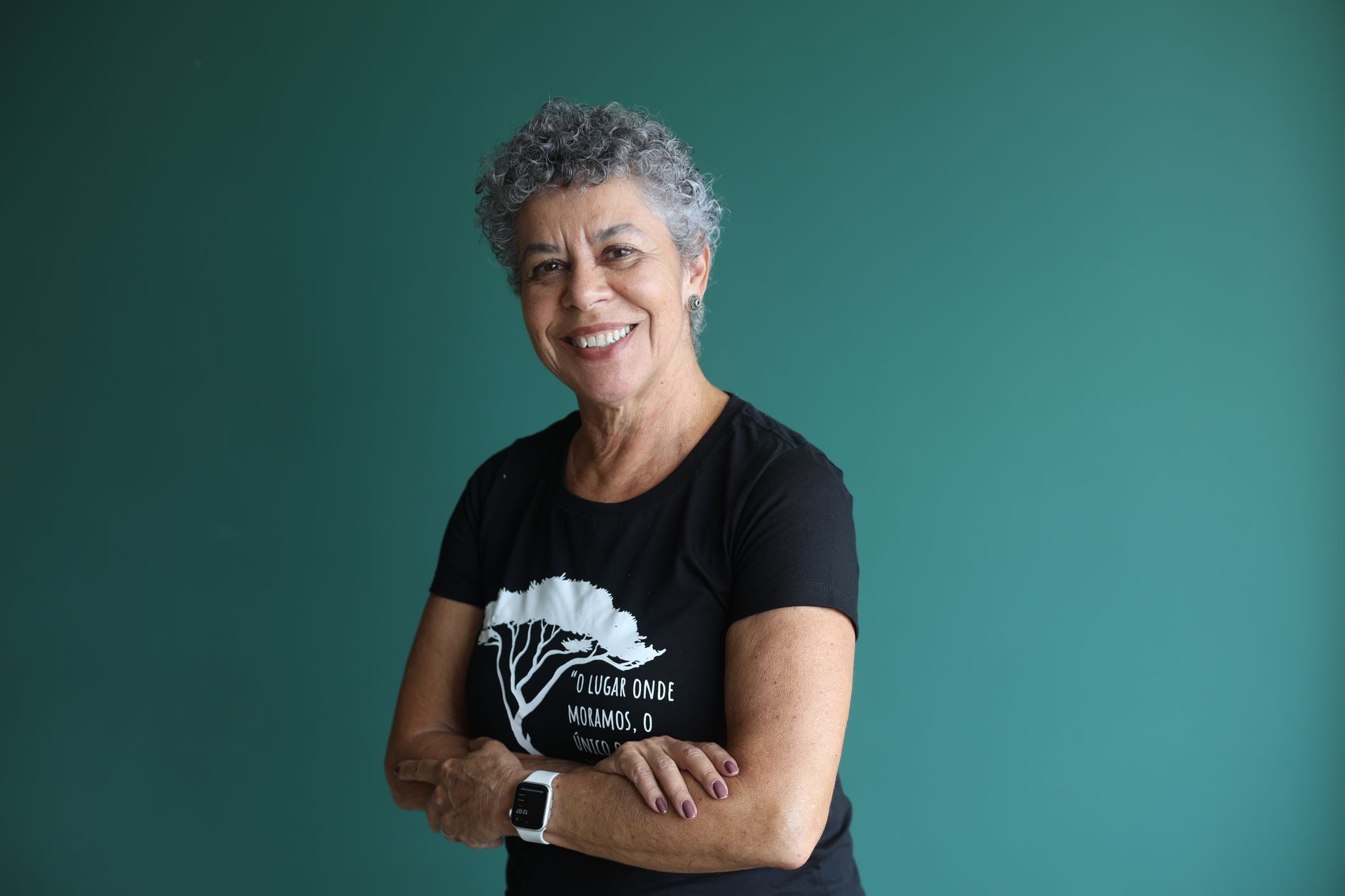 Cristina Pinho, vestindo camiseta preta, em pé com os braços cruzados contra um fundo azul-petróleo.