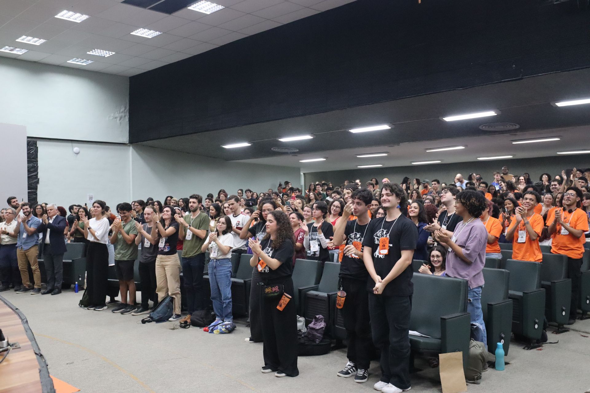 Grande público de pé aplaudindo em um salão de conferências, muitos vestindo camisetas laranja.