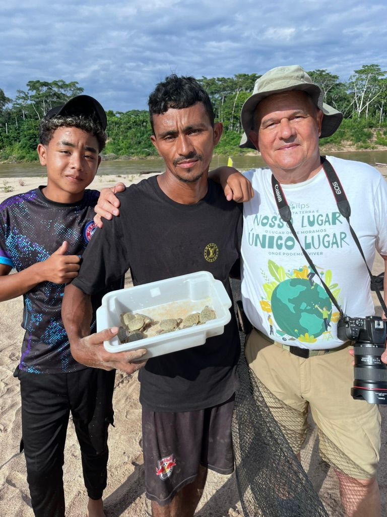 Three people standing outdoors, smiling, holding a plastic container of fish near a river or shore.