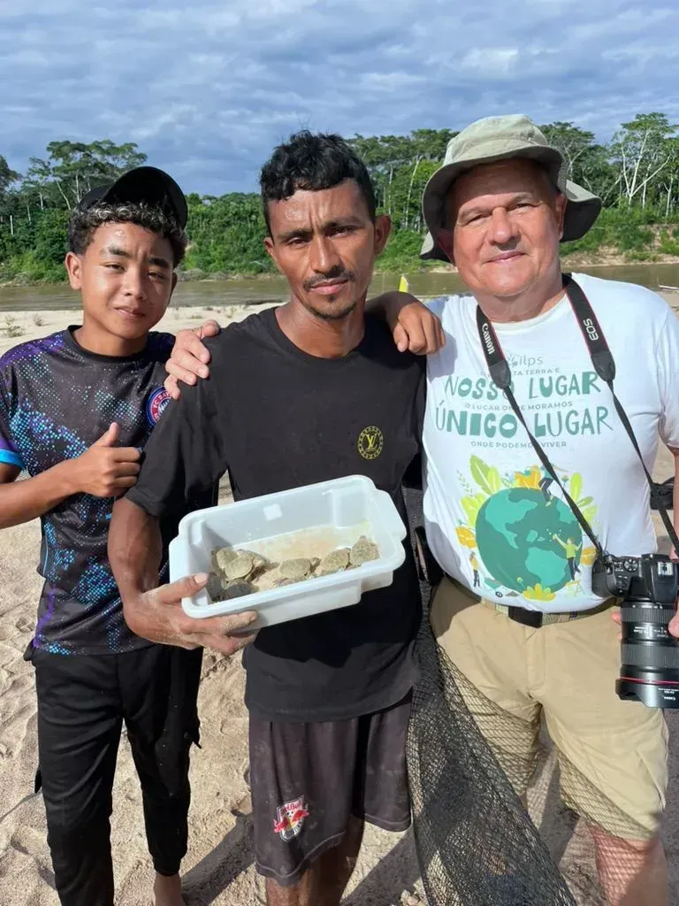 Three people standing outdoors, smiling, holding a plastic container of fish near a river or shore.