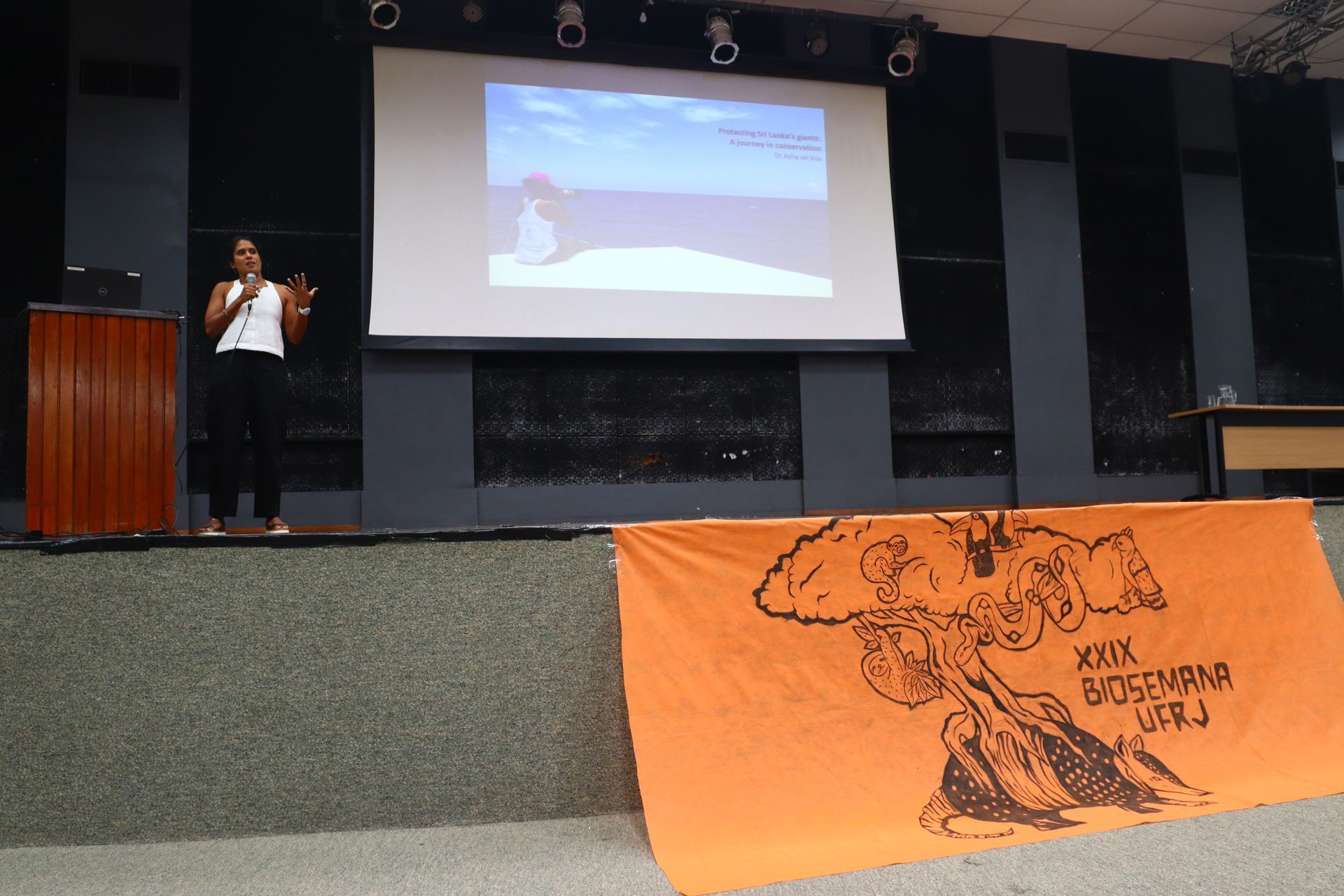 Pessoa falando em um palco ao lado de uma tela de projeção, com uma faixa laranja estendida na frente.
