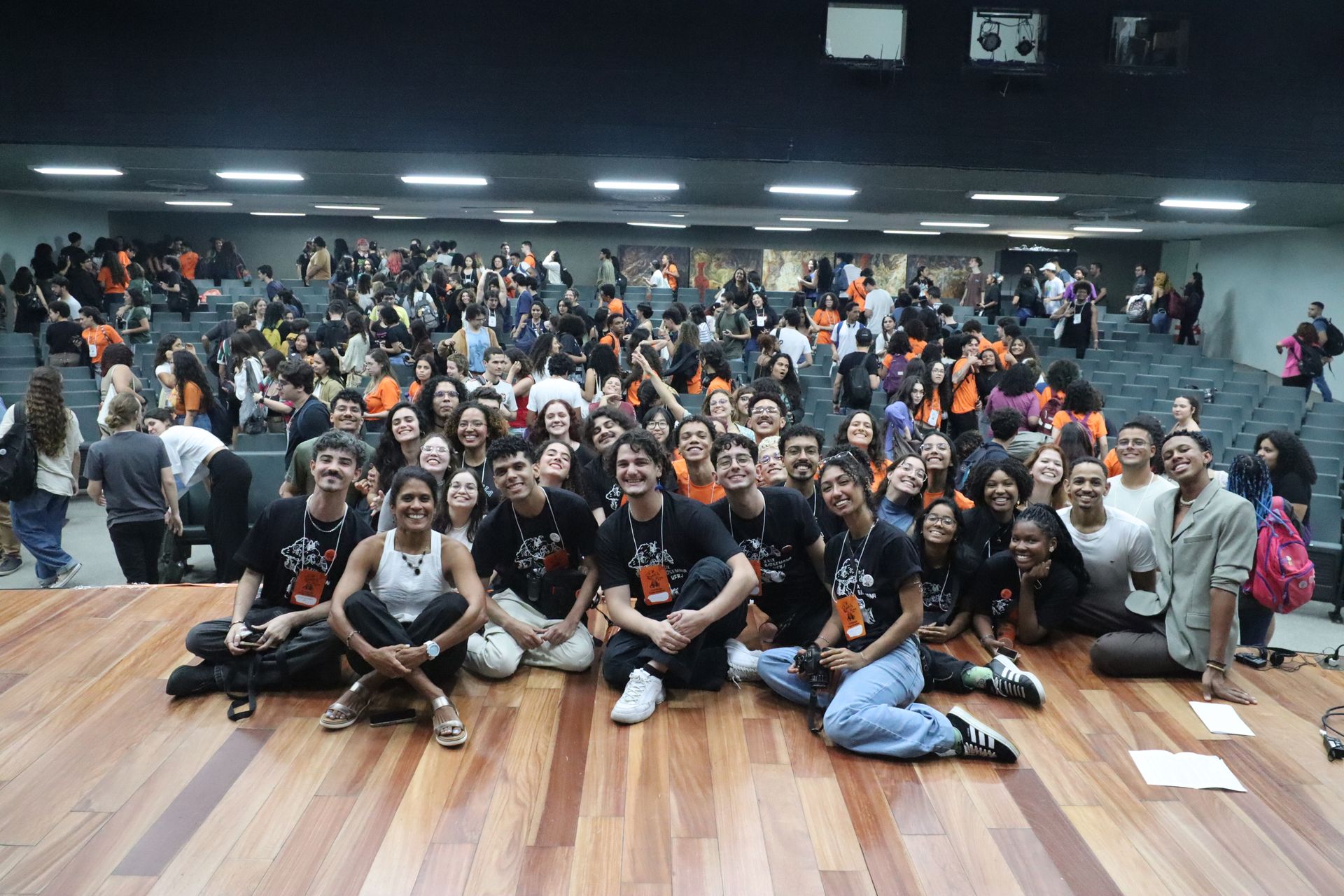 Um grande grupo posando em um piso de madeira em uma academia ou estúdio, com luzes brilhantes e uma parede escura ao fundo.