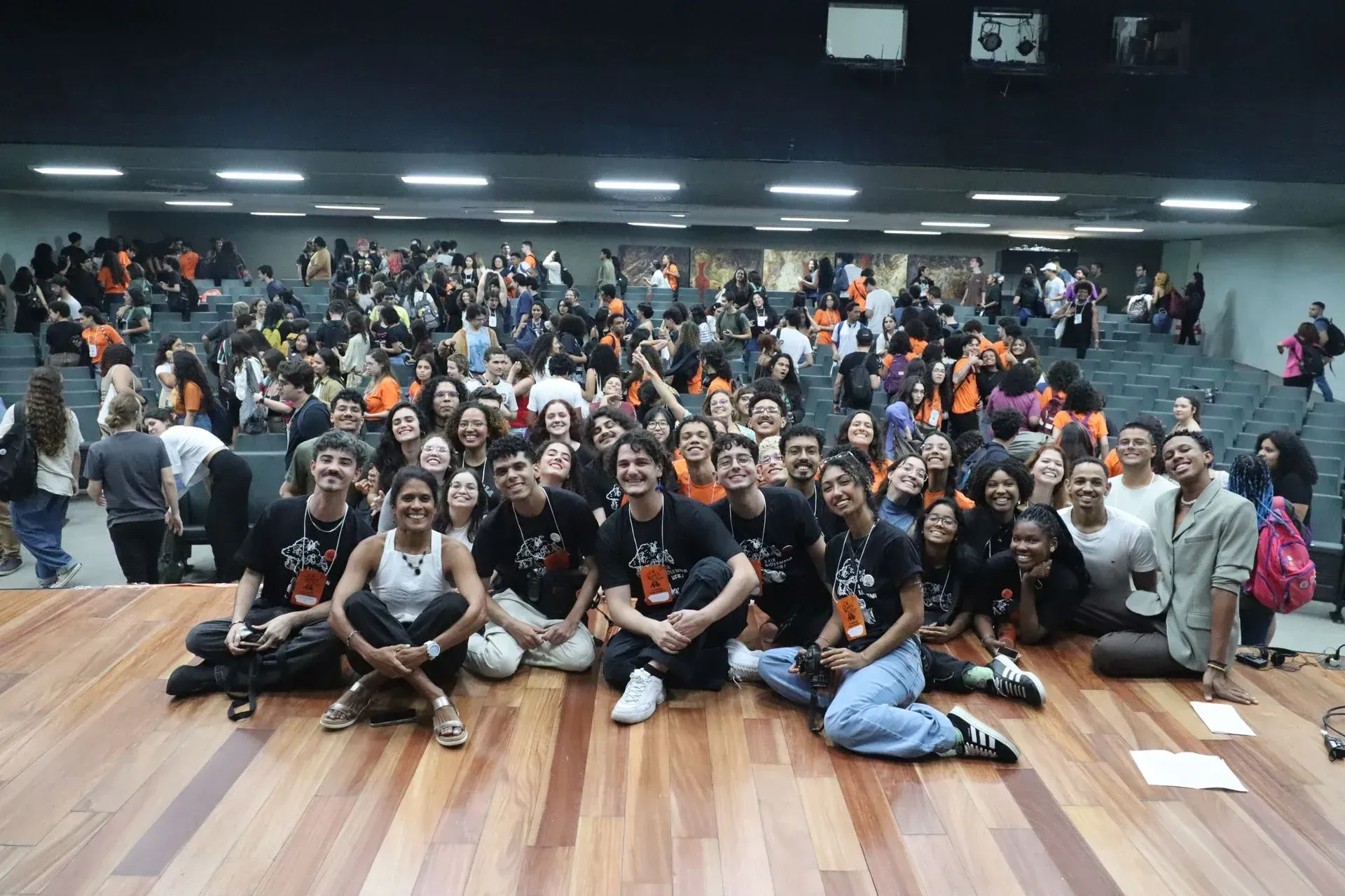 Um grande grupo posando em um piso de madeira em uma academia ou estúdio, com luzes brilhantes e uma parede escura ao fundo.