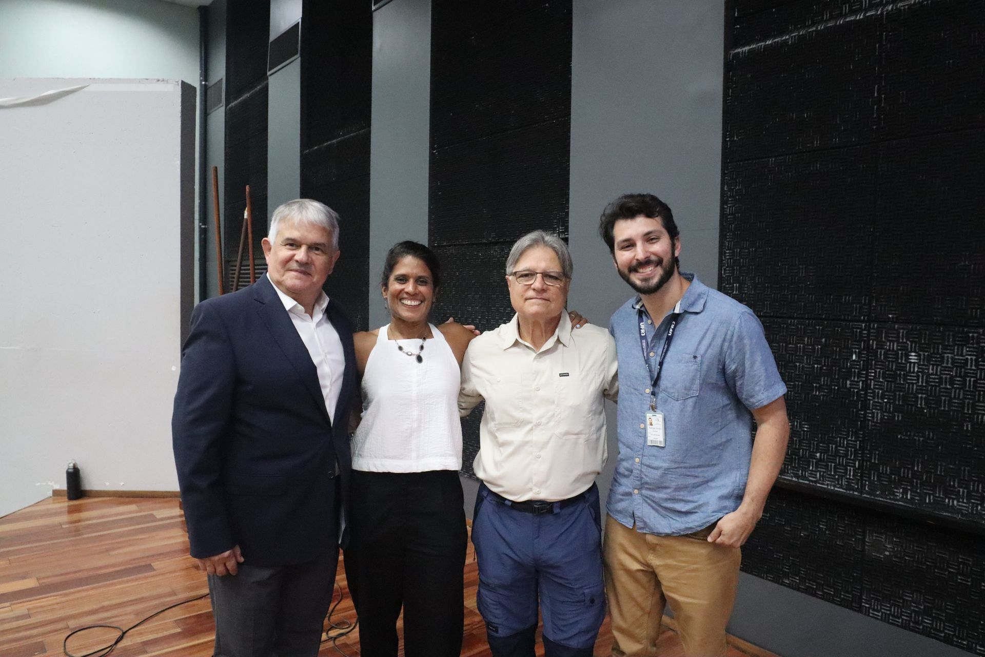 Quatro pessoas posando em um palco dentro de casa, com fundo preto e piso de madeira polida.