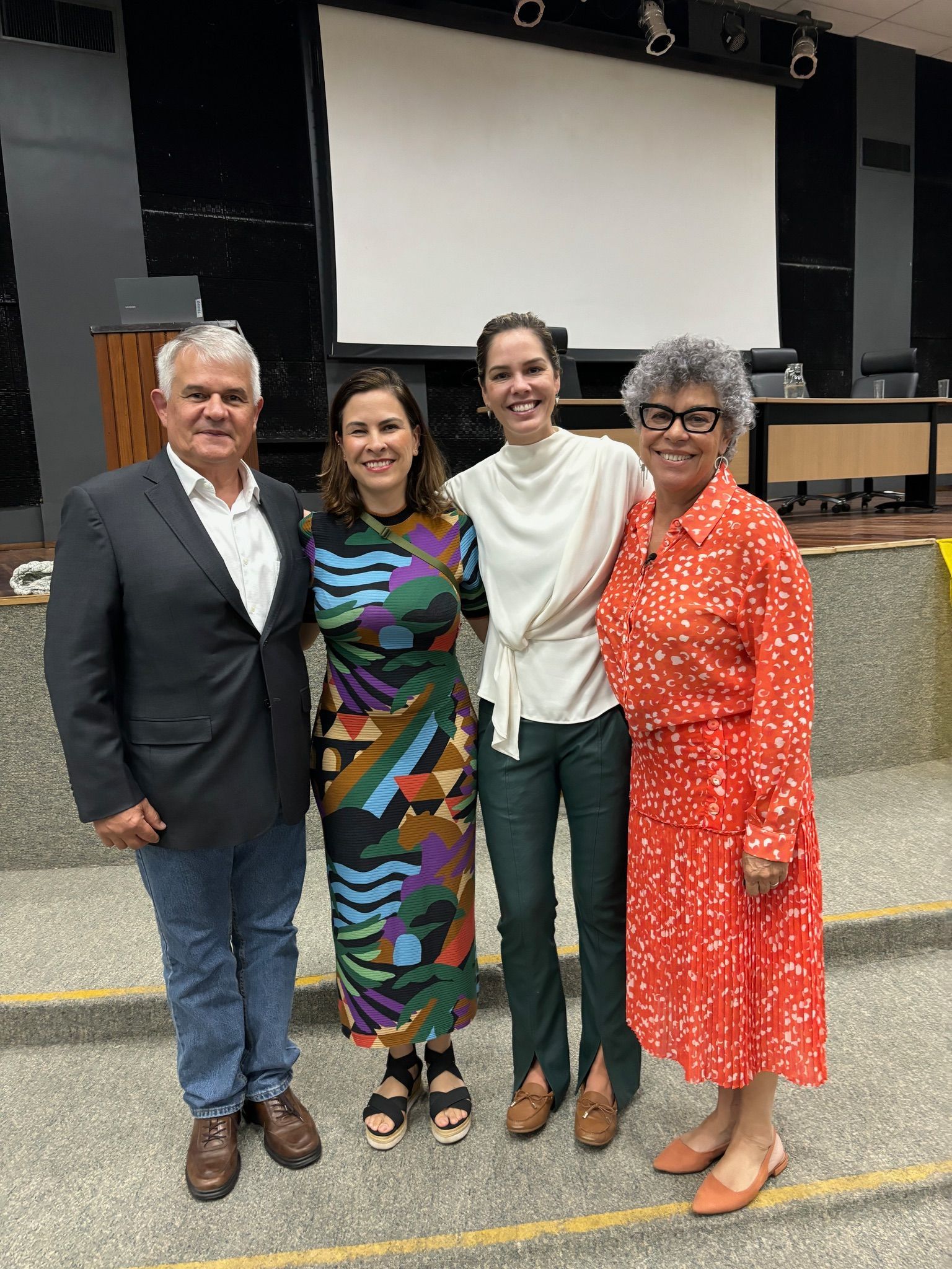 Quatro pessoas juntas nos degraus de um auditório. Duas mulheres e dois homens. O cenário tem uma tela e alguns móveis.