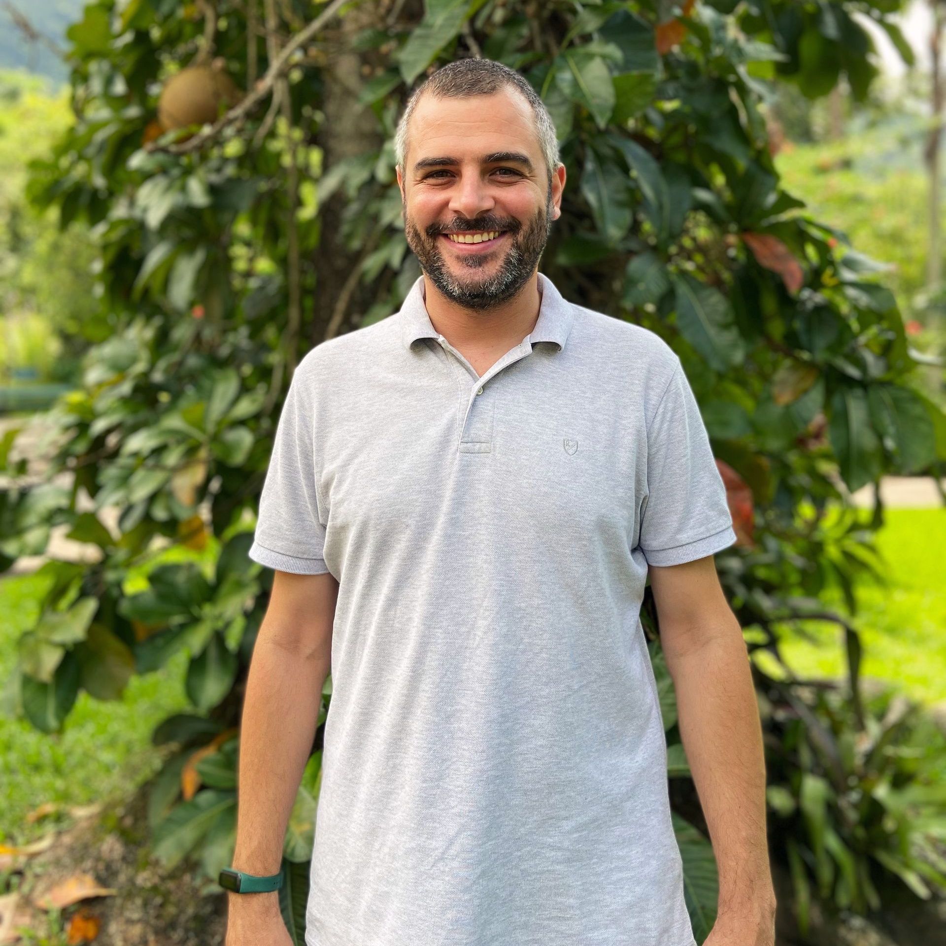 Um homem com barba vestindo uma camisa pólo cinza está parado em frente a uma árvore.