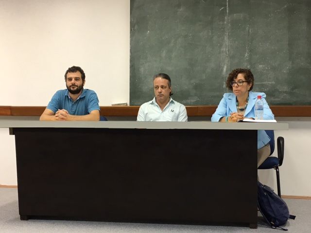 Três pessoas estão sentadas em uma mesa em frente a um quadro negro.