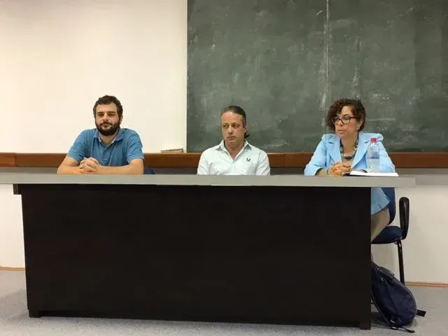 Três pessoas estão sentadas em uma mesa em frente a um quadro negro.