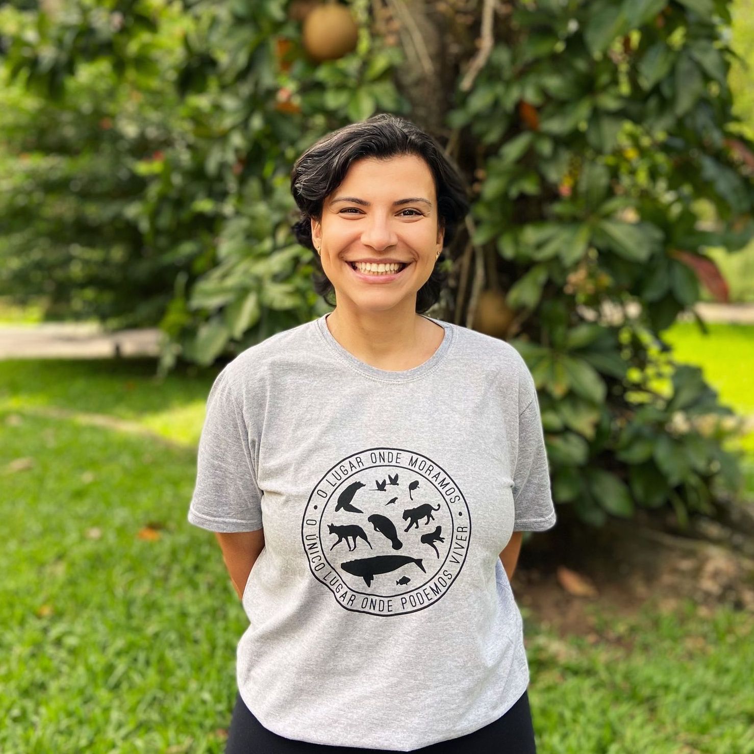 Uma mulher vestindo uma camiseta cinza está parada em frente a uma árvore.