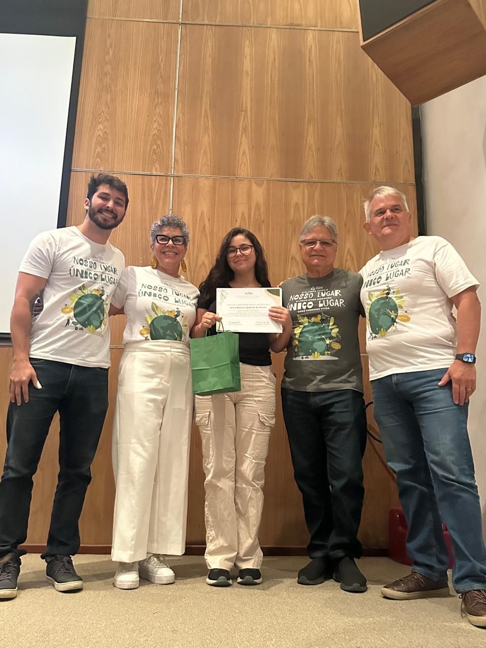 Cinco pessoas no palco seguram um cheque gigante, em pé diante de uma parede com painéis de madeira.