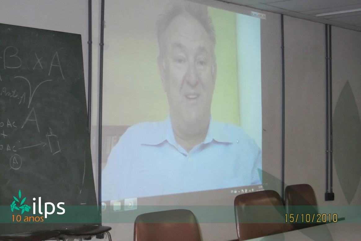 O rosto de um homem é projetado em uma tela em frente a um quadro-negro que diz ilps