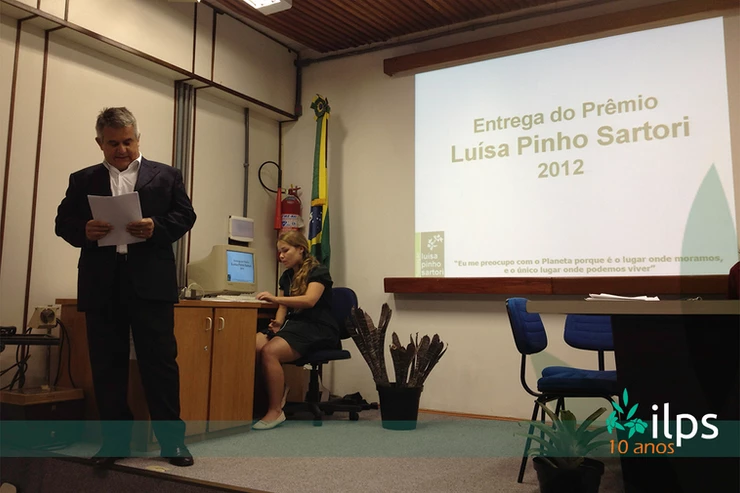 Um homem diante de uma tela de projetor que diz entrega do prêmio luisa pinho sartori 2012