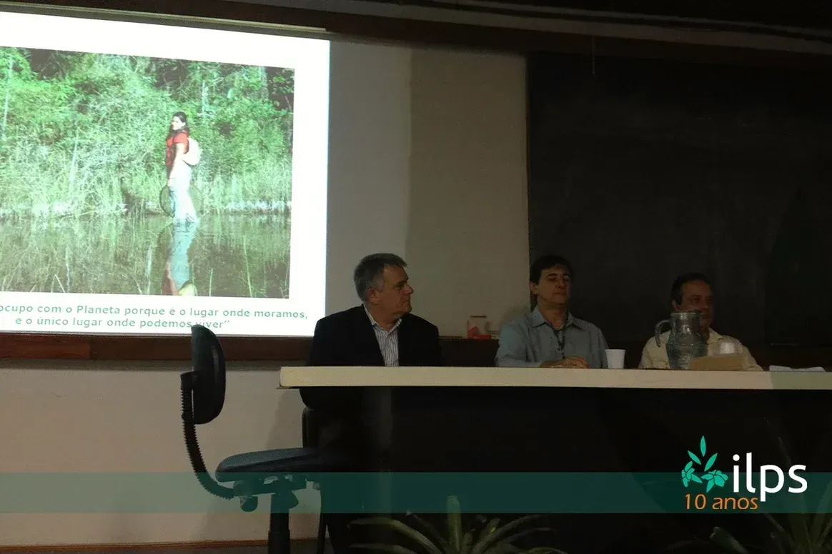 Dois homens estão sentados a uma mesa em frente a uma tela de projetor.
