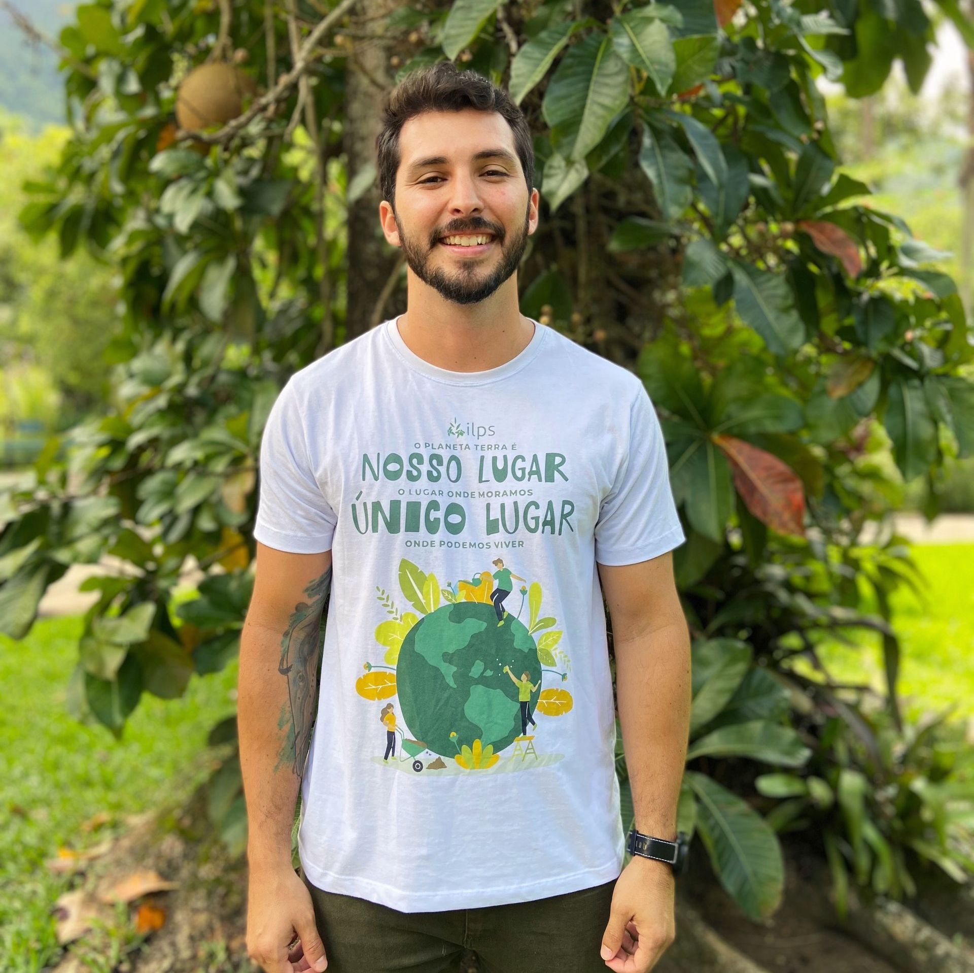 Um homem vestindo uma camiseta branca com um globo verde está parado em frente a uma árvore.