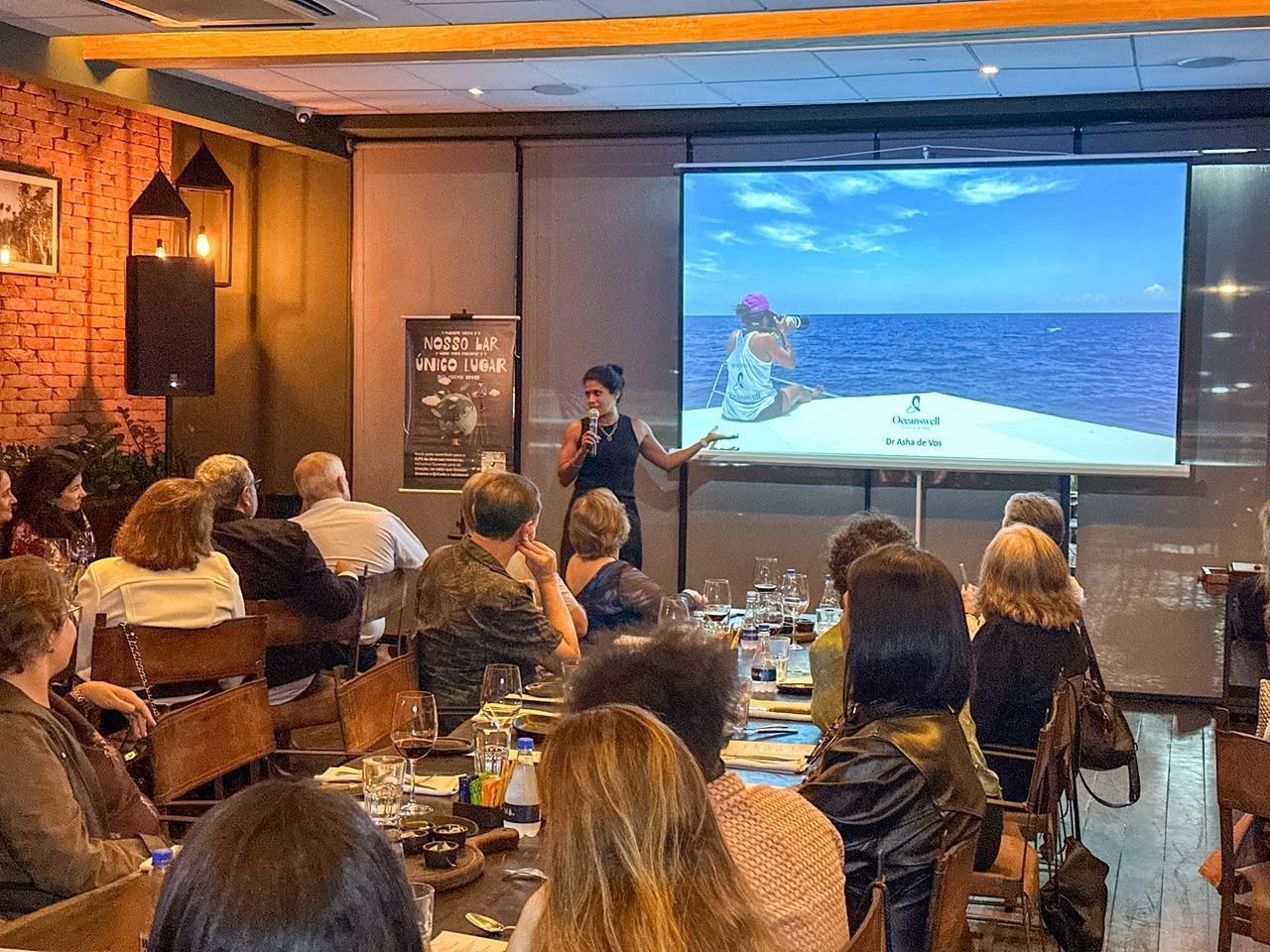 Audience watches a presentation in a restaurant, with a speaker beside a large screen showing a seaside scene.