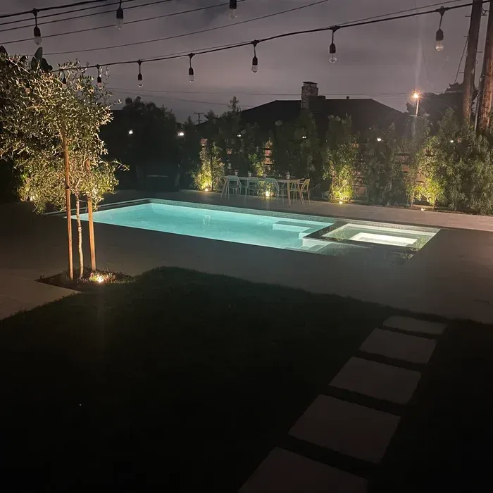 Nighttime backyard pool with turquoise water, string lights overhead, and a lit pathway through the lawn