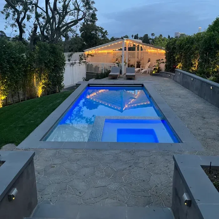 Backyard pool glowing blue at dusk, with a lit patio and pergola in the background.