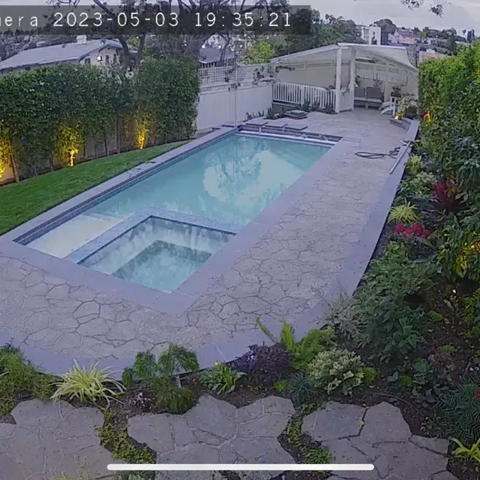 Backyard pool and hot tub beside a patio, with greenery and a covered seating area.