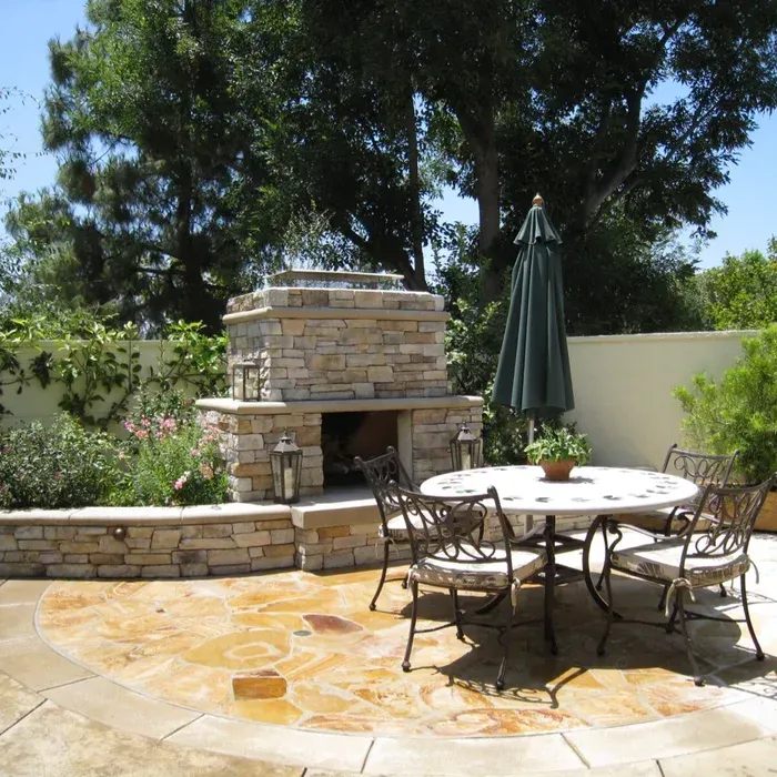 Patio with stone outdoor fireplace, round table, chairs, and a closed umbrella in a sunny garden