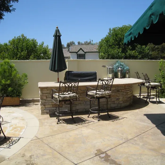 Sunny backyard patio with stone bar, metal chairs, and a closed umbrella under a blue sky