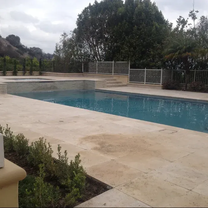 Outdoor swimming pool with blue water, beige deck, and trees under an overcast sky.