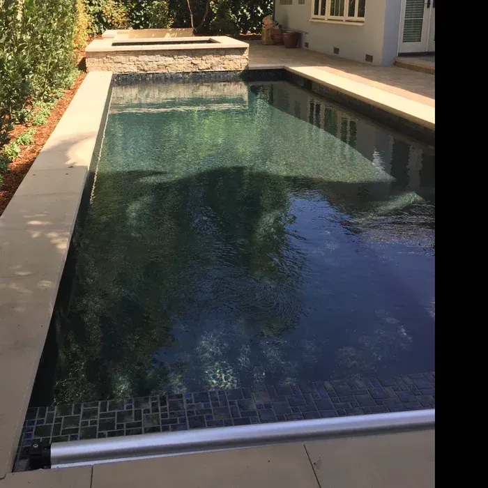 Rectangular backyard swimming pool with dark water, stone deck, and house patio in sunlight