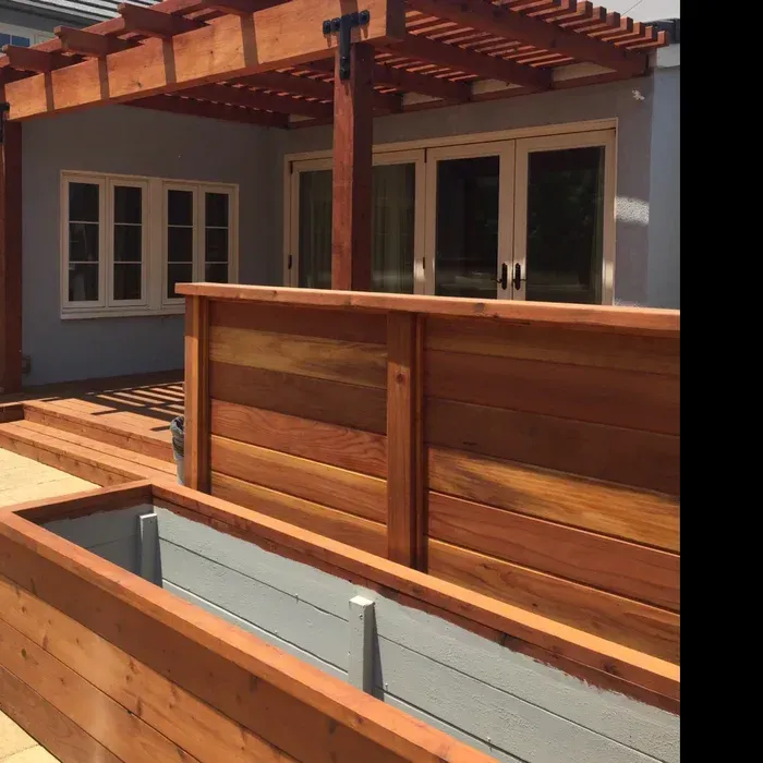 Wooden deck with built-in benches and pergola outside a gray house with white-framed doors and windows