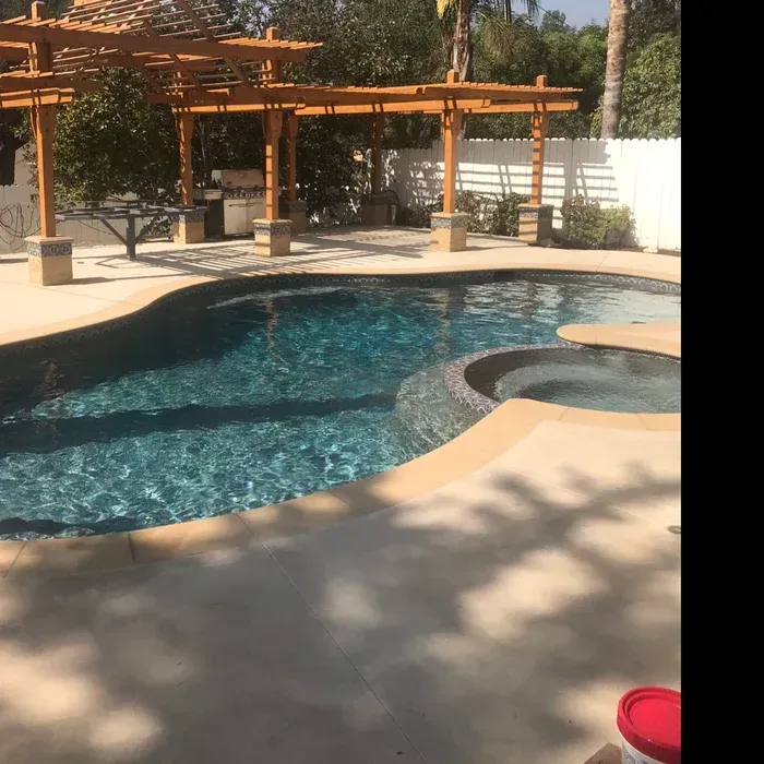 Backyard pool with curved blue water, hot tub, and wooden pergola in sunny patio area