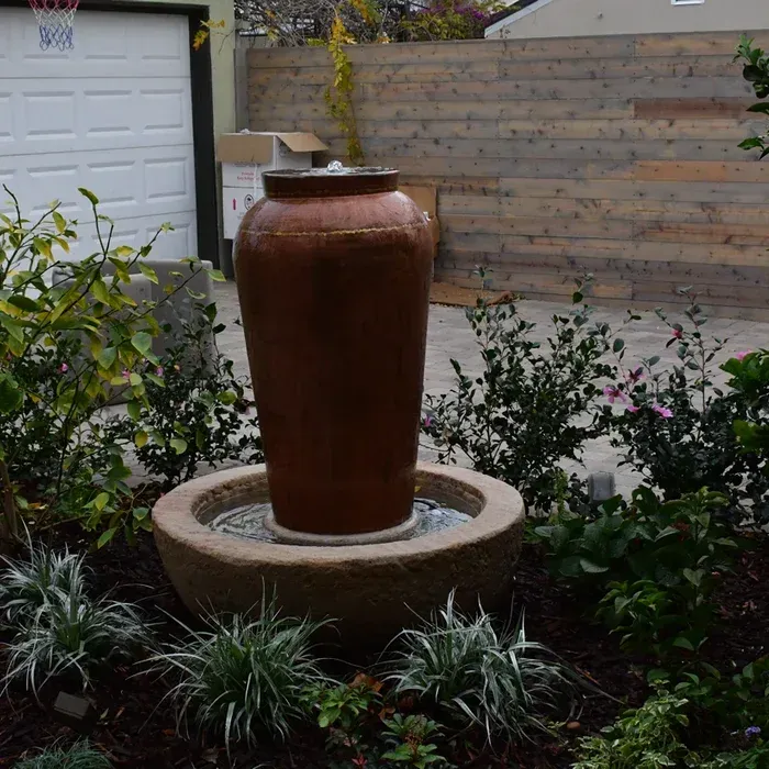 Brown ceramic fountain in a landscaped garden bed with white garage and wooden fence in the background