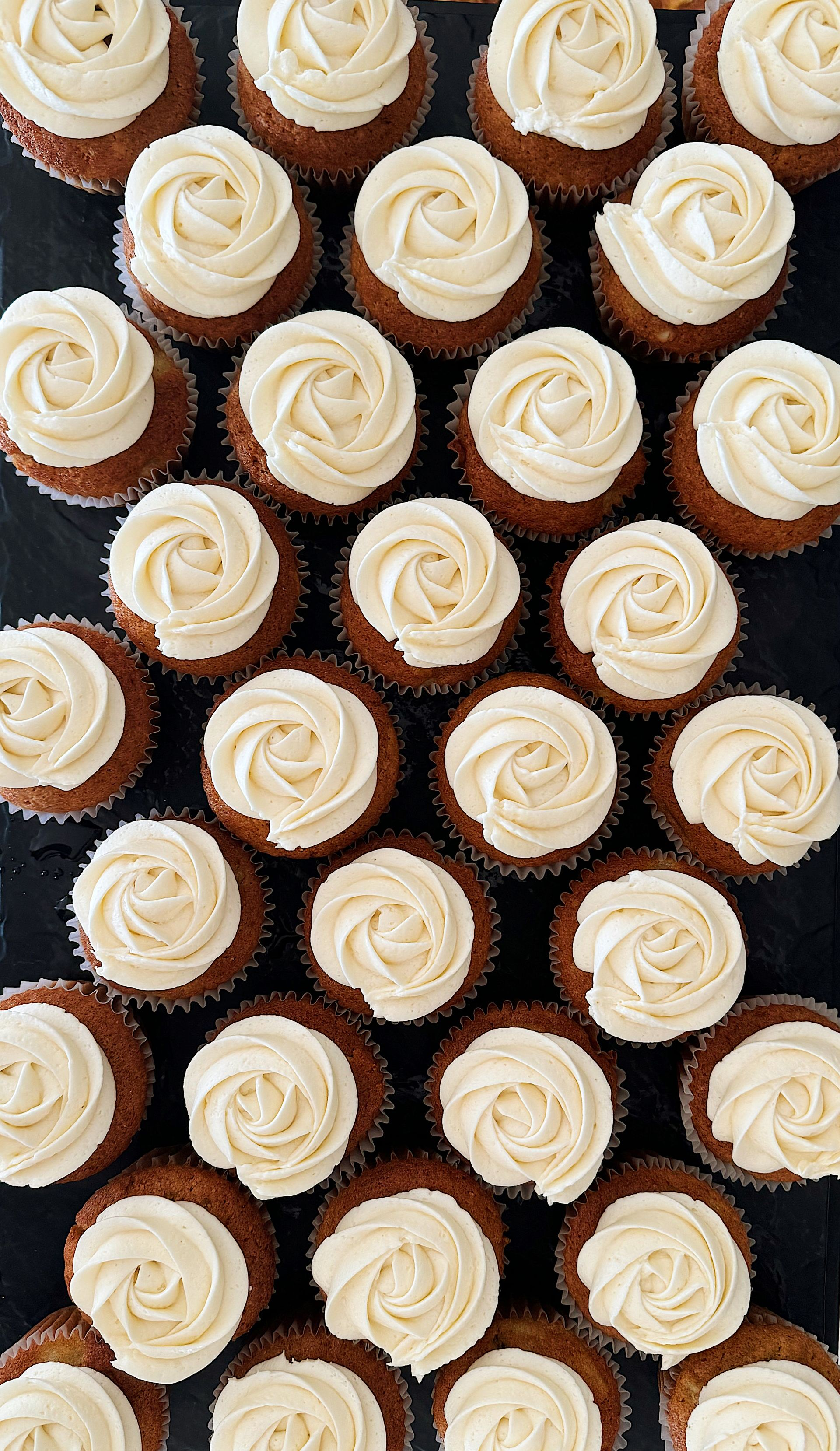 A top-down view of many small cupcakes arranged in a grid, topped with white frosting swirls on a dark background.