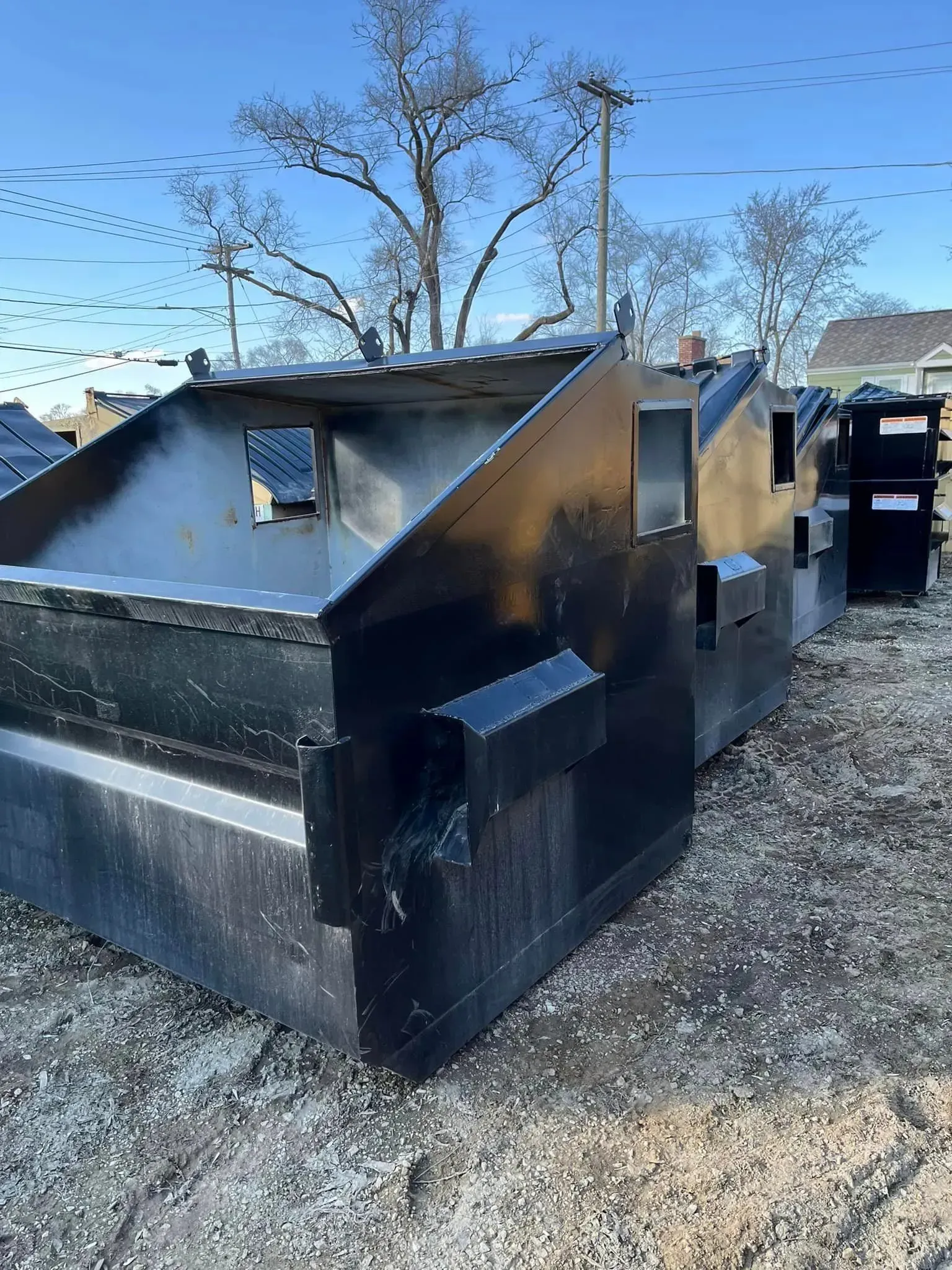 Several black metal waste dumpsters lined up on a gravel lot under a clear blue sky.