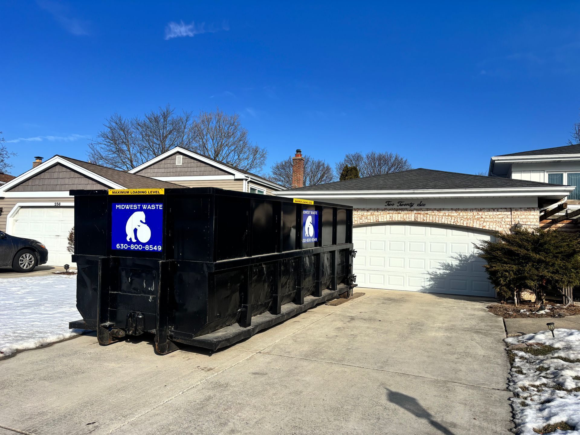 A black roll-off dumpster sits on a residential concrete driveway in front of a house on a sunny day.