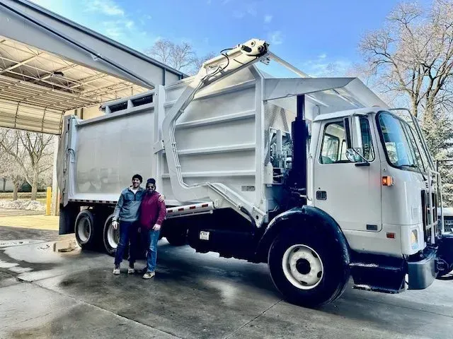 Two people stand in front of a large white garbage truck parked under a covered structure on a sunny day.