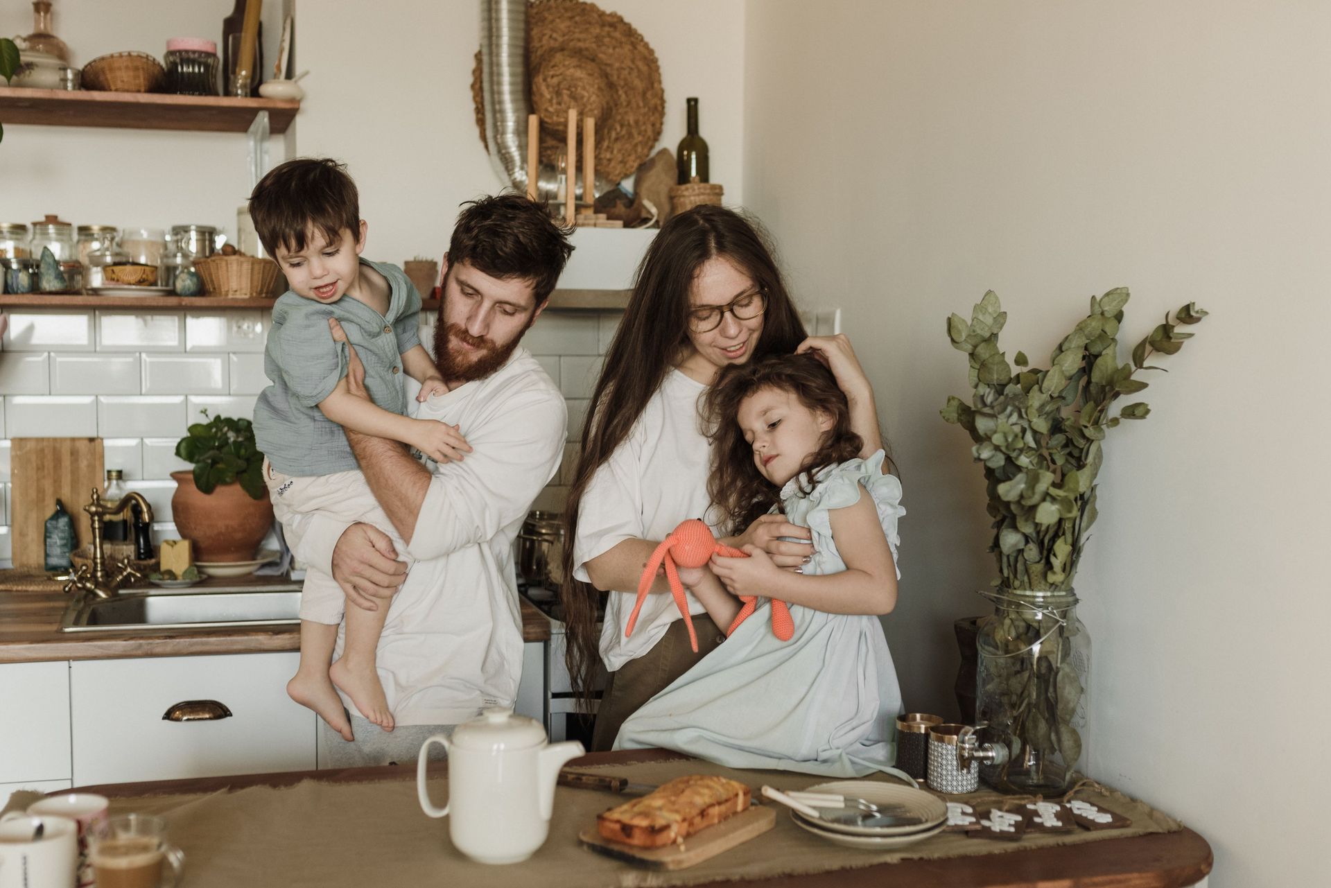 Family in a cozy kitchen, with two children and two adults gathered around a table with food and mugs.