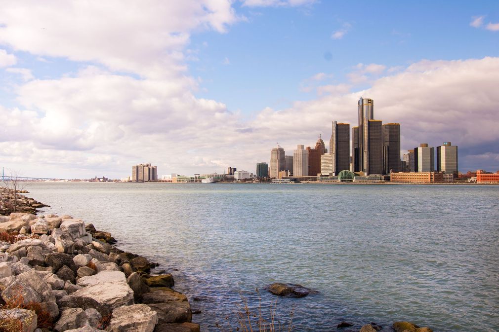 Rocky shoreline beside calm water with a city skyline under a partly cloudy sky