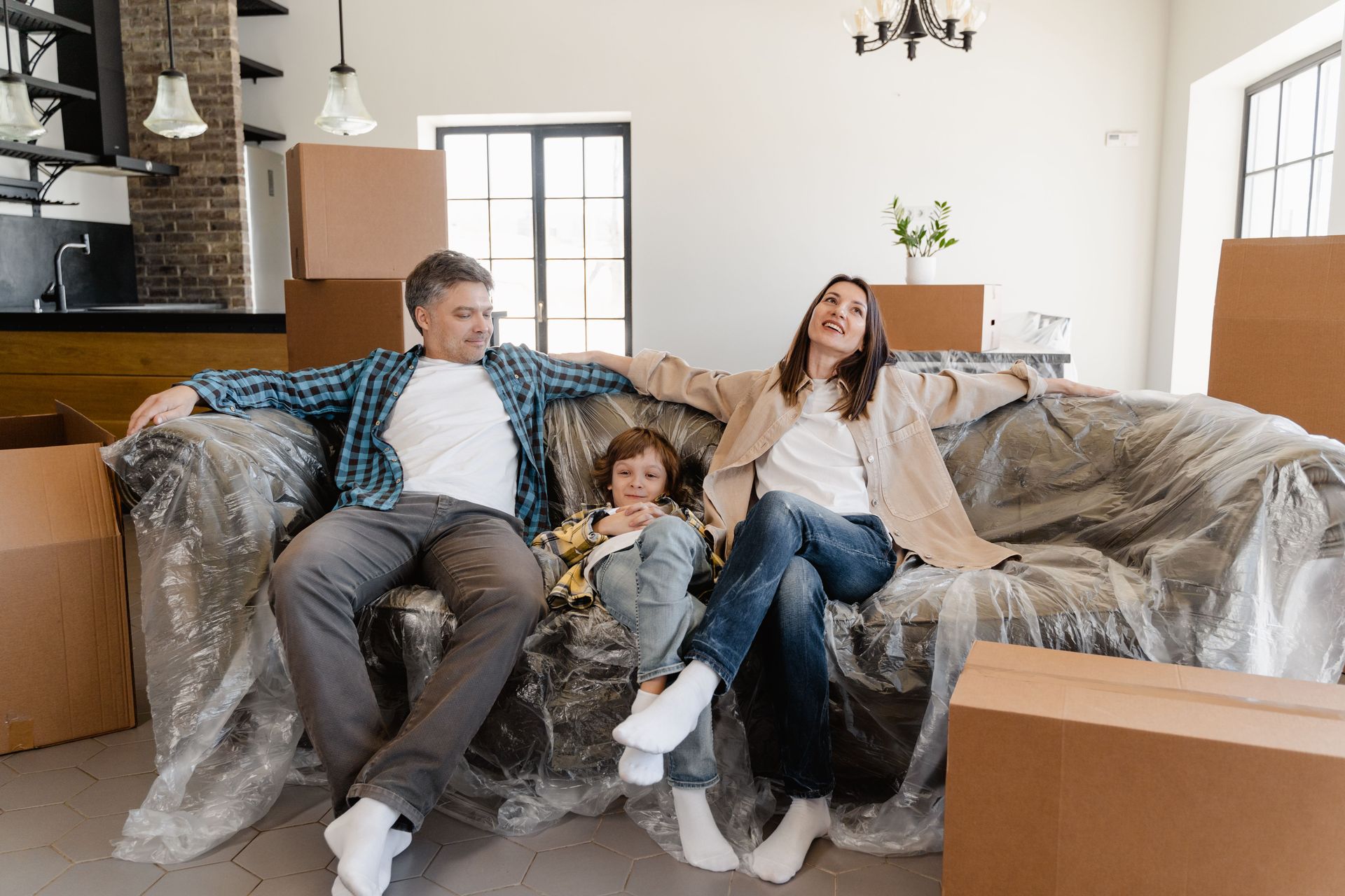 Three people relaxing on a couch in a bright living room surrounded by moving boxes.