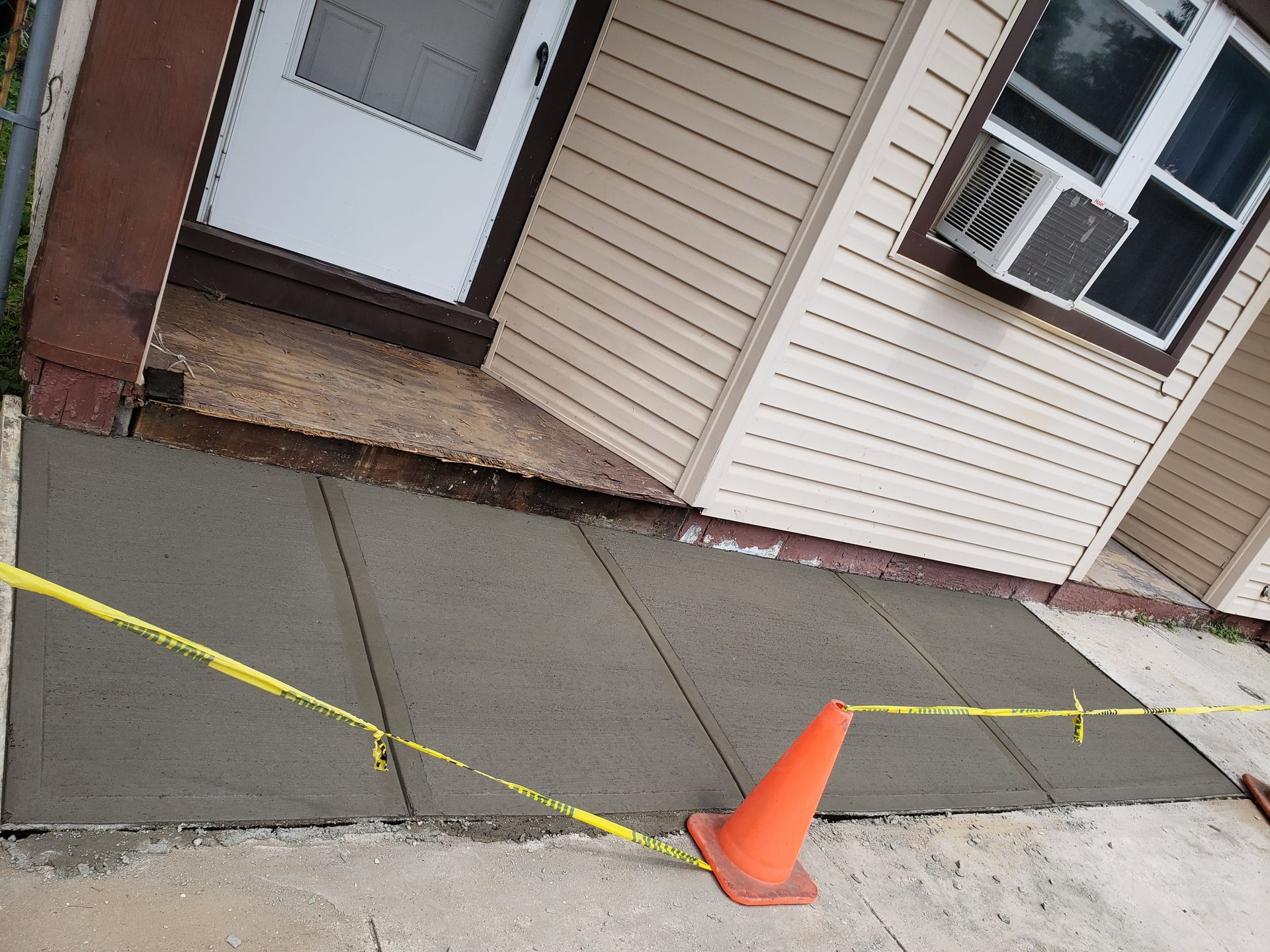 Tape cordons off a cracked sidewalk beside a building, with an orange cone near a doorway.