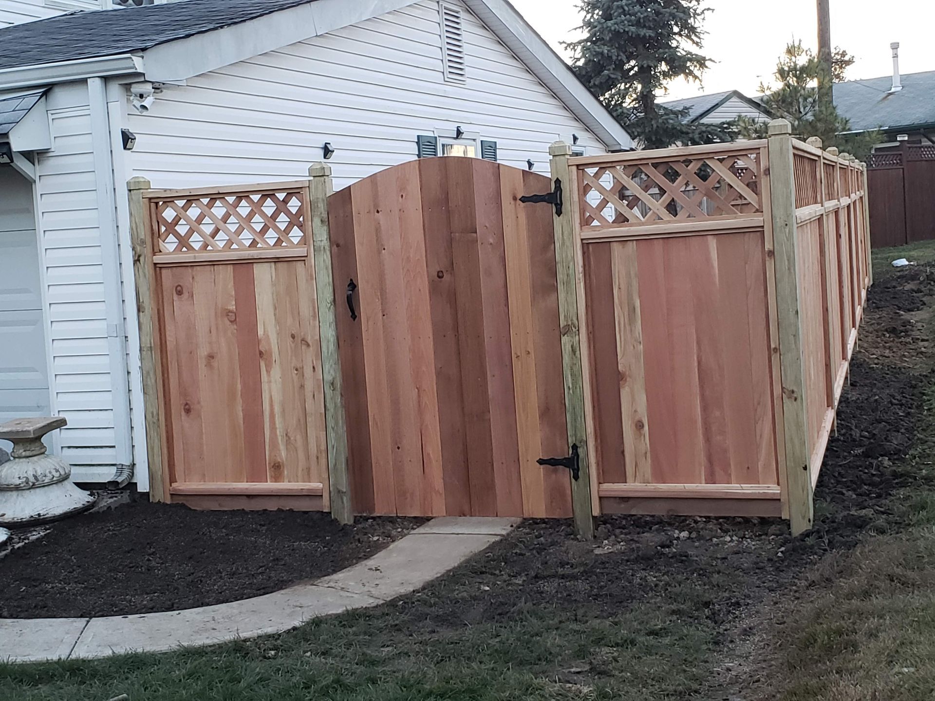 Wooden backyard gate and fence beside a white house, with lattice tops and a curved path leading to it.