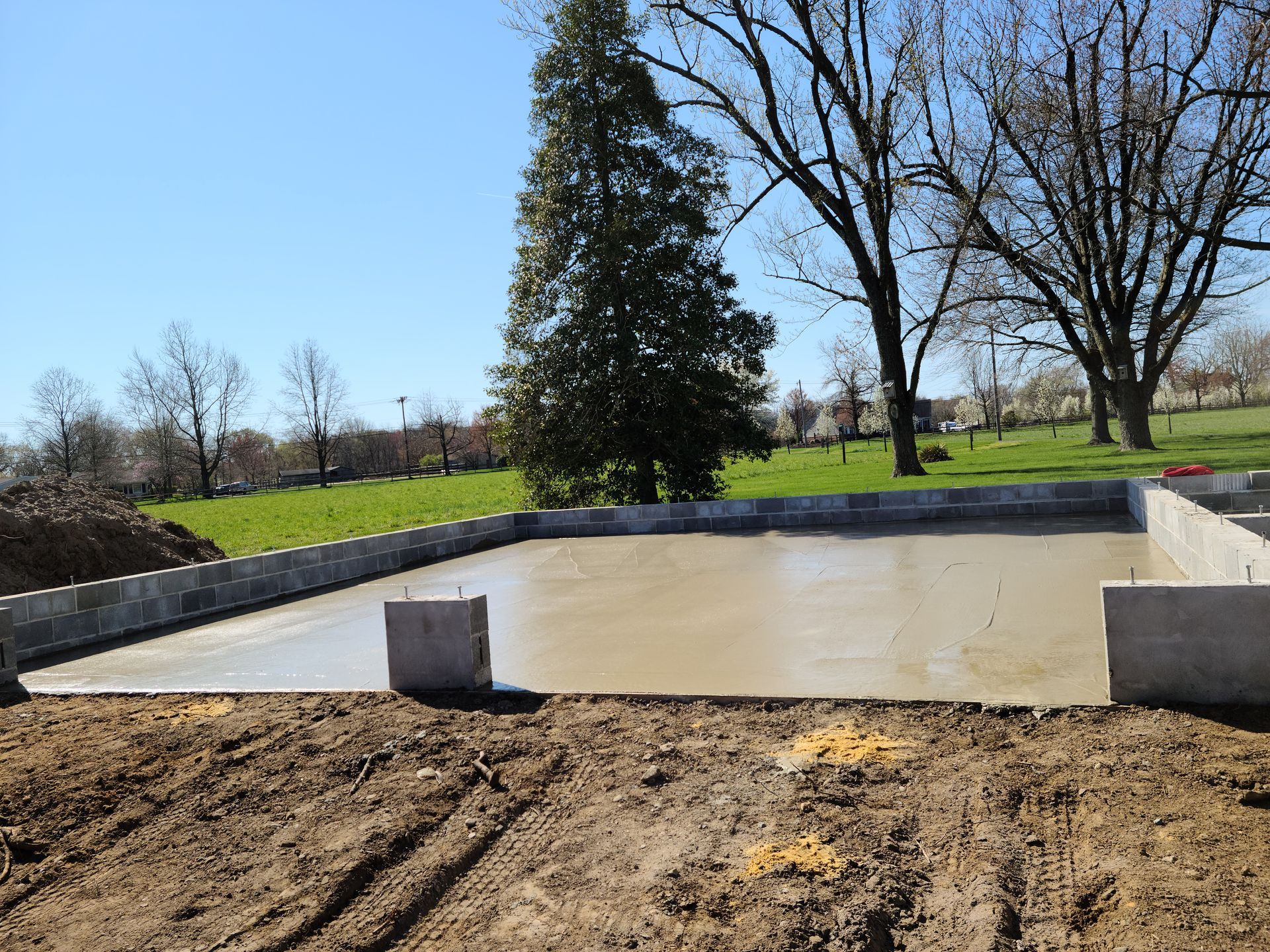 Concrete foundation slab in a wooded backyard, with dirt foreground and trees under a clear sky.