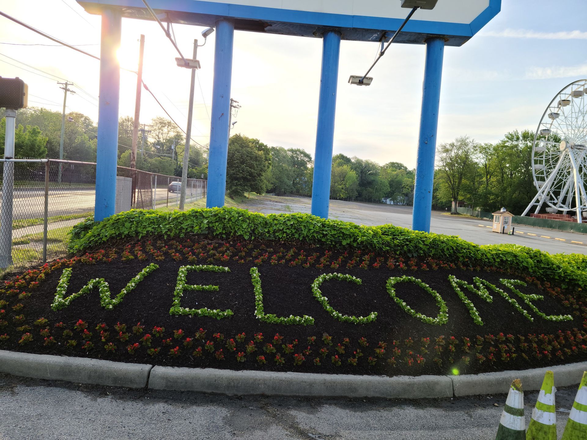 Flower bed with “WELCOME” in green plants at an amusement park entrance, with blue supports and rides in the background