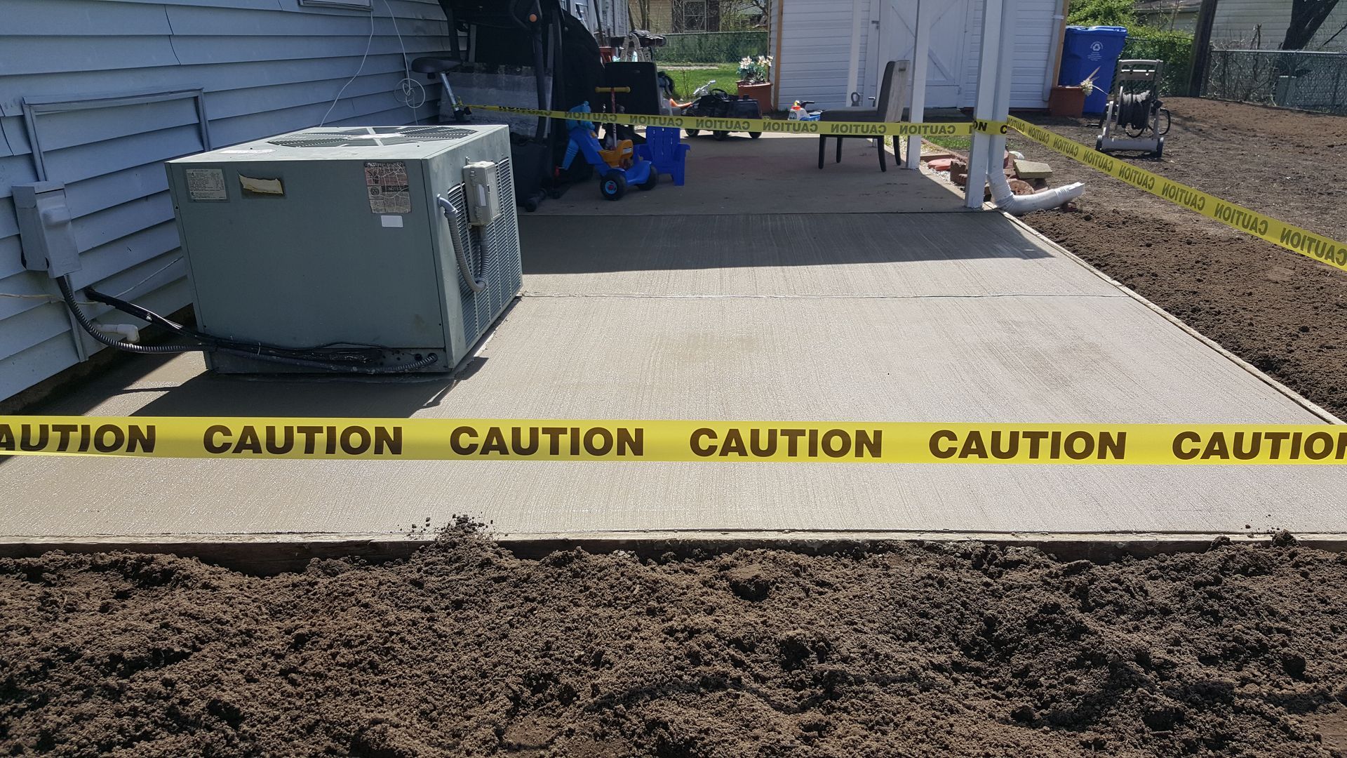 Caution tape blocks a paved area beside a house, with an outdoor unit and worksite materials nearby.