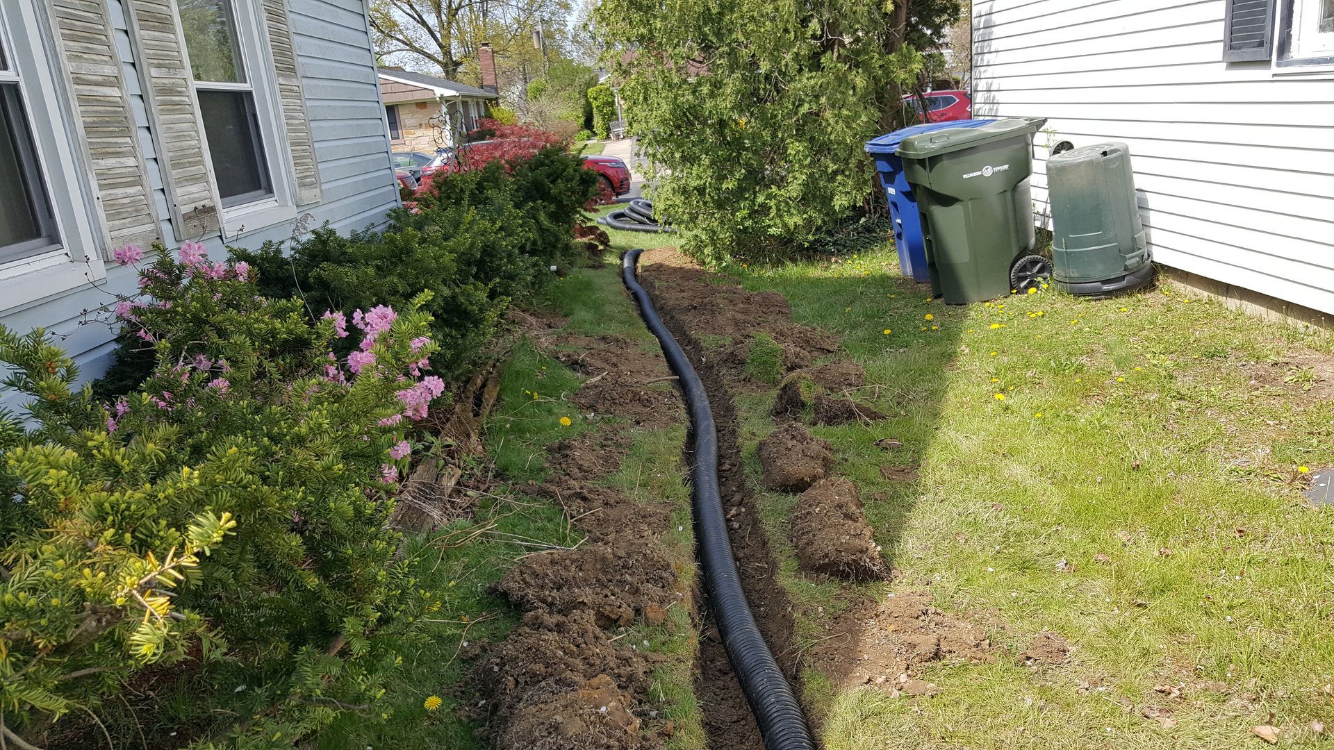 Side yard with a black hose along a flower bed beside a house, green lawn, shrubs, and trash bins nearby
